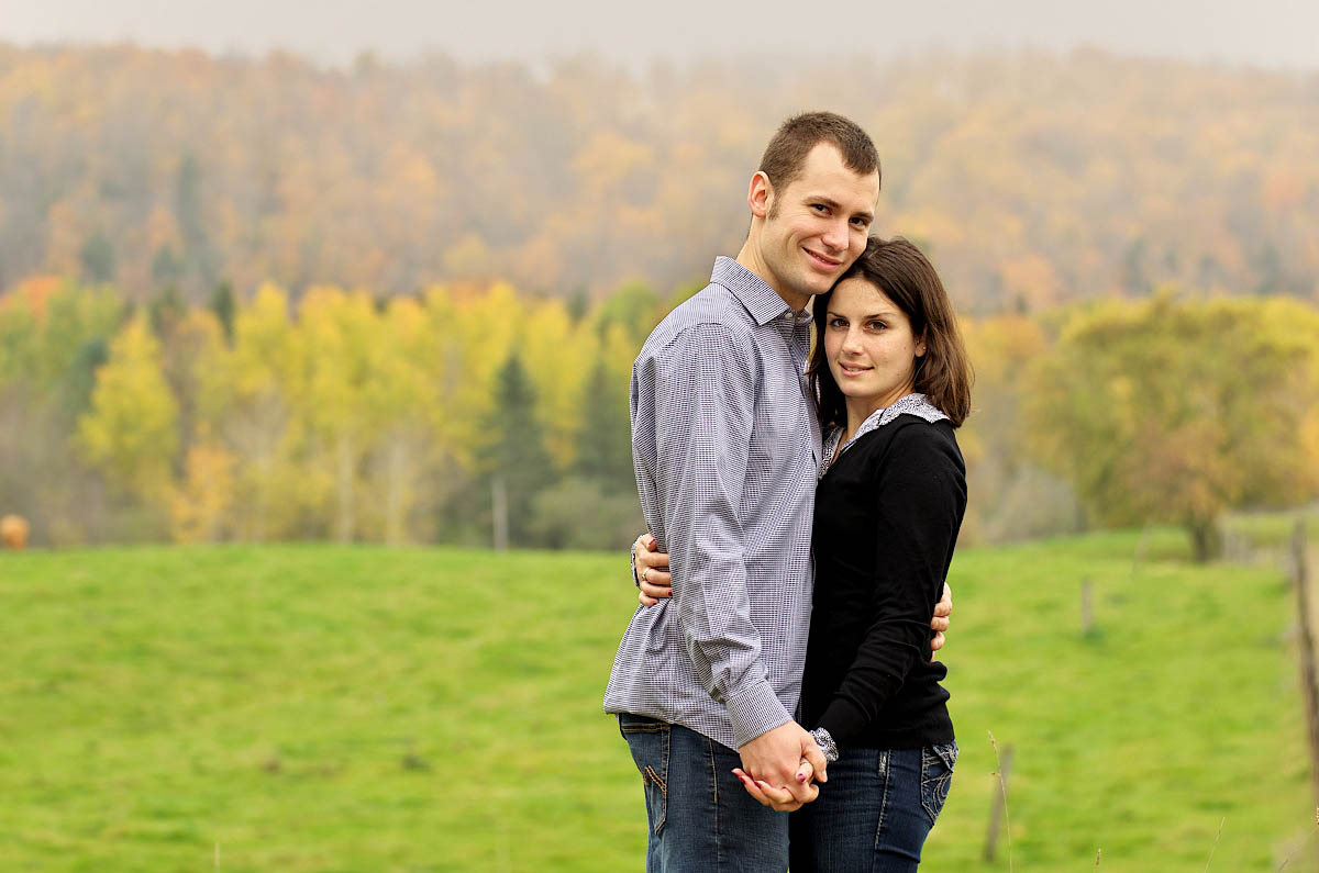 Young couple in farm field with fall colours and valley behind them during Omemee engagement session near Peterborough