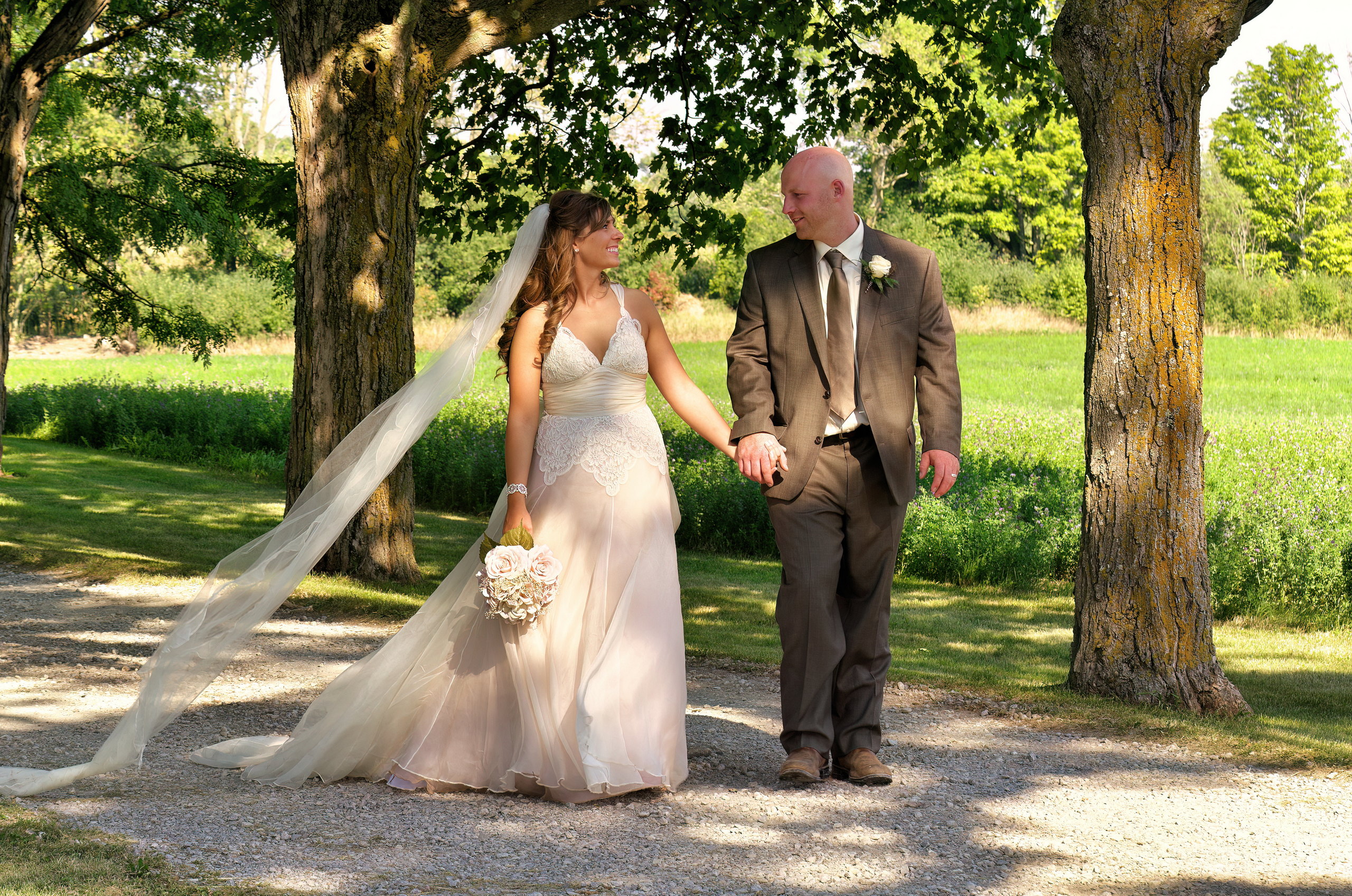 Bride and Groom walk hand in hand down a shady driveway at an Omemee farm wedding